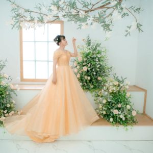 A bride in a gown poses elegantly amidst lush floral arrangements indoors.